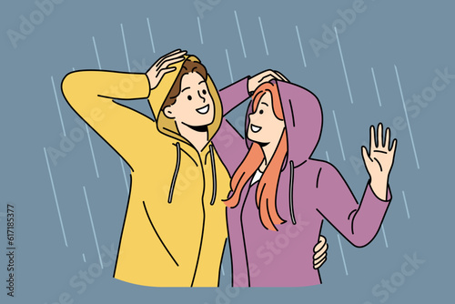 Happy couple stands in rain in waterproof hoods and hugs rejoicing at cloudy weather and first autumn downpour. Cheerful man and woman walking in rain during date or having romantic conversation