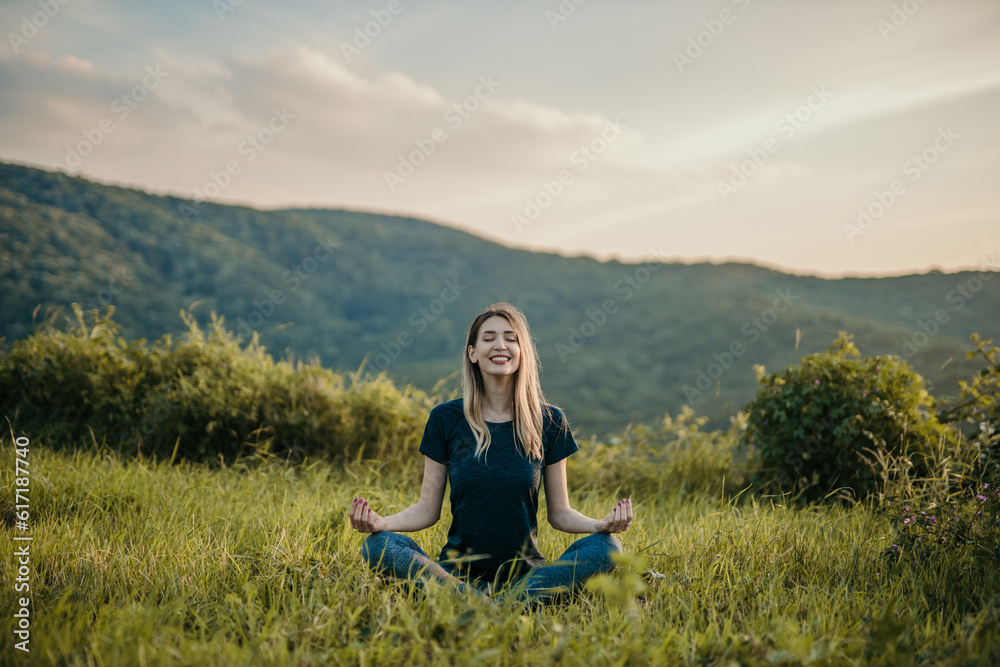 The rising sun casts a warm glow on a woman as she peacefully sits in a lotus position, finding tranquility in nature.