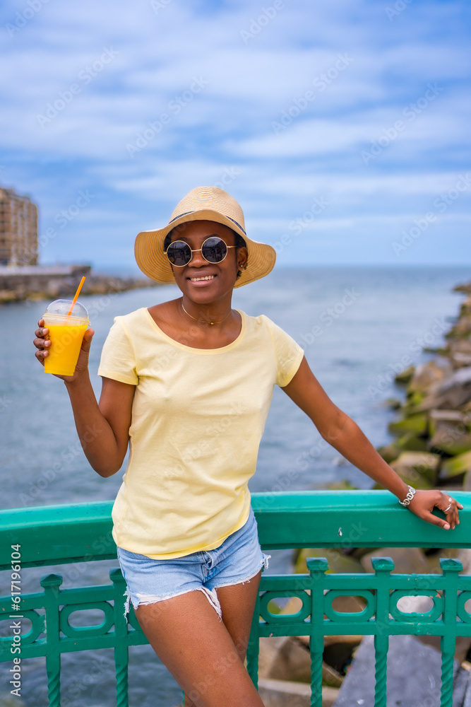 Black ethnic woman having an orange juice by the sea, tourist enjoying the summer
