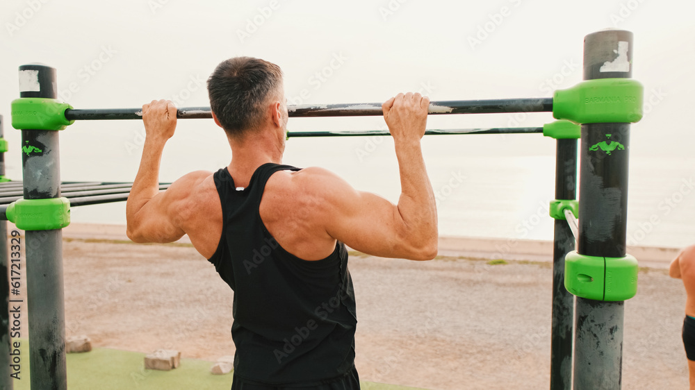 Naklejka premium Middle-aged muscular man in sportswear pulls himself up on the horizontal bar on sea background, Back view
