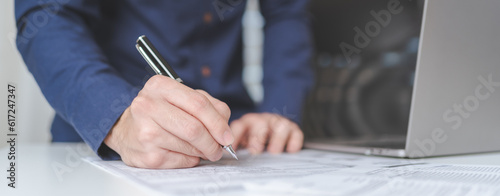 Businessman hand holds pen, writing on business paperwork for accept agreement contract on business investment project. Young adult business man signing on financial document deal. Copy space. Banner.