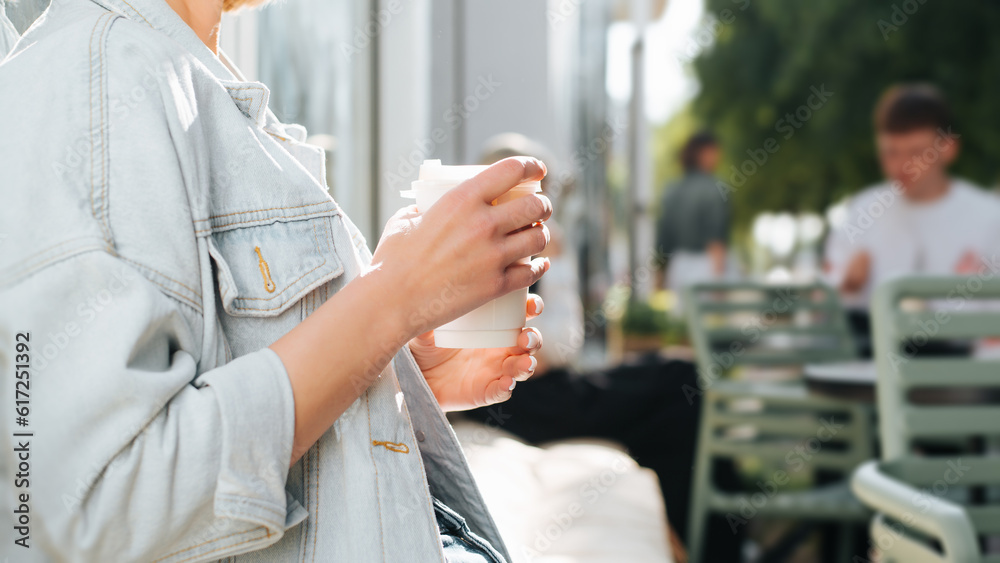 Close-up of caucasian female hand holding white disposable cup of coffee while sitting in street cafe on sunny summer day. Side view of unrecognizable woman relaxing outdoors, lifestyle