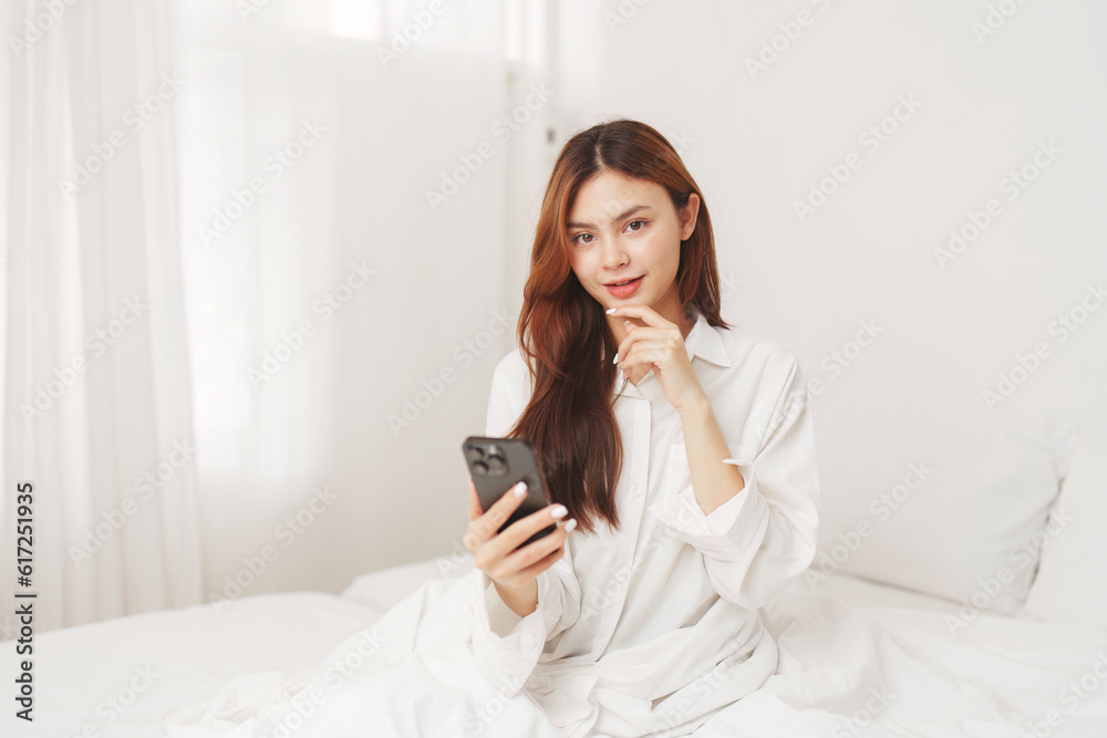 Happy smiling asian woman stretching on bed in the morning and holding mobile phone
