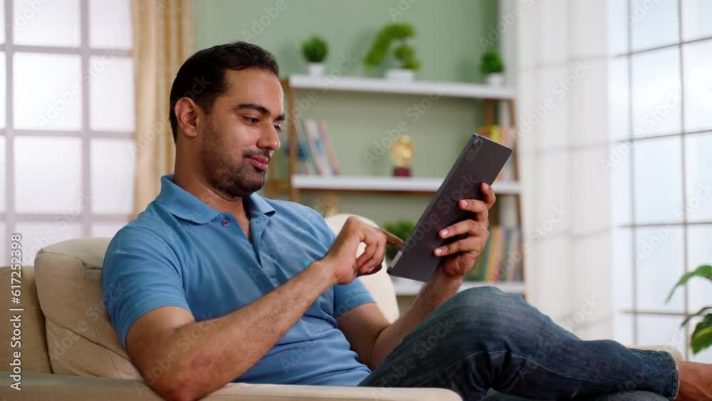 Happy relaxed indian man using digital tablet while sitting on sofa at home - concept of leisure time, modern lifestyle and surfing web. Happy relaxed indian man using digital tablet while sitting on sofa at home - concept of leisure time, modern lifestyle and surfing web.