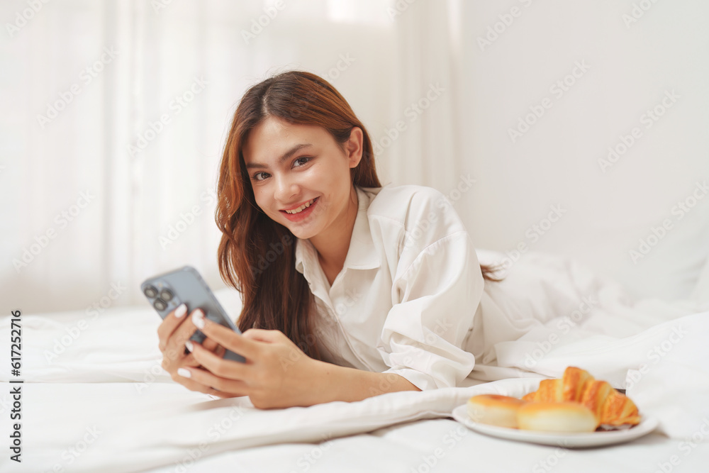 Happy smiling asian woman stretching on bed in the morning and holding mobile phone Happy smiling asian woman stretching on bed in the morning and holding mobile phone