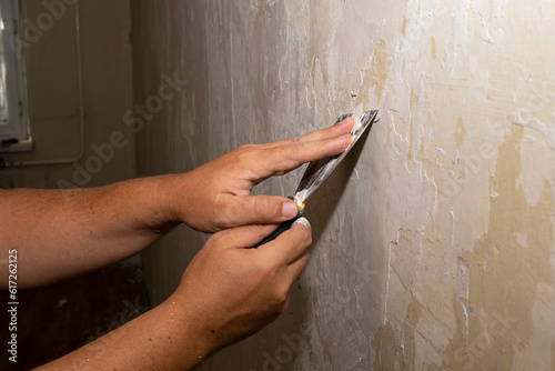 A spatula in men's hands removes old plaster from the wall in the house. The concept of cleaning walls.