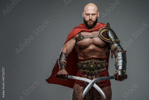 Bald and bearded gladiator poses standing tall with two swords while wearing light armor. This fearsome and imposing warrior emanates power and strength against a neutral gray background