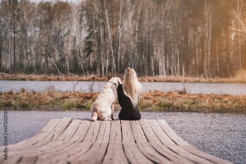 Girl with a dog on the bridge near the lake. White labrador on a walk with a woman near the river. sitting with their backs