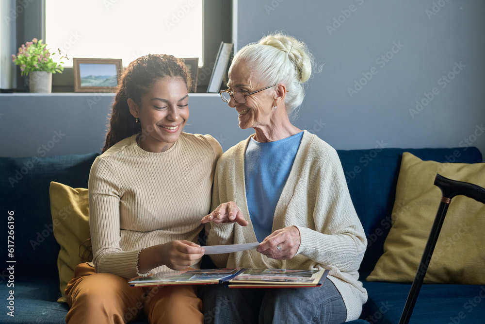 © AnnaStills - Happy old woman in eyeglasses looking at her granddaughter while sitting on sofa next to her and showing her album with family photos © AnnaStills - Happy old woman in eyeglasses looking at her granddaughter while sitting on sofa next to her and showing her album with family photos