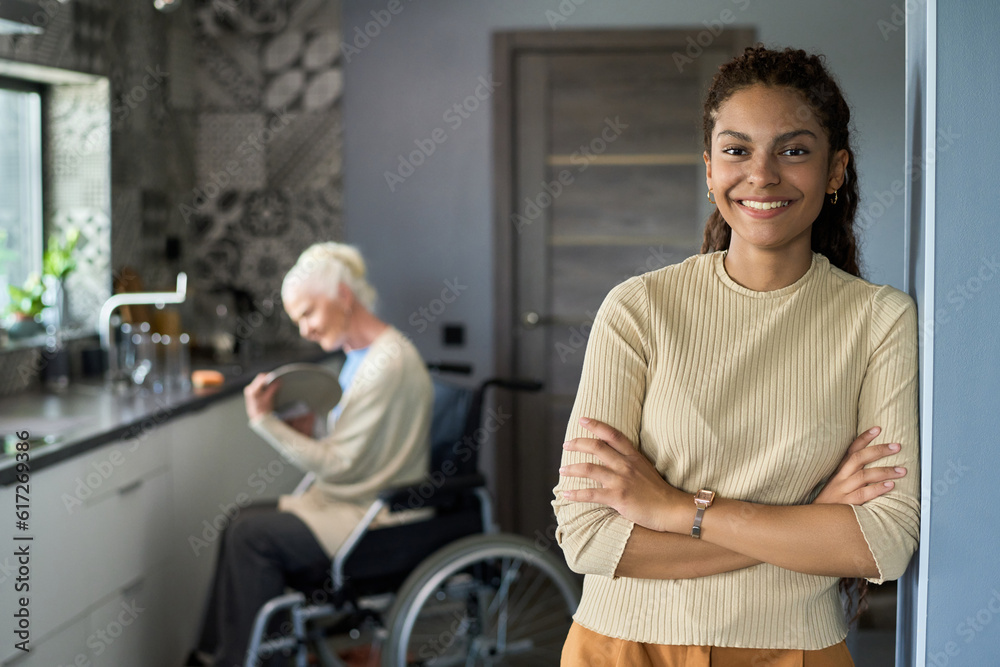 Young cheerful multiethnic teenage girl looking at camera while standing against her grandmother with disability wiping plate in the kitchen Young cheerful multiethnic teenage girl looking at camera while standing against her grandmother with disability wiping plate in the kitchen