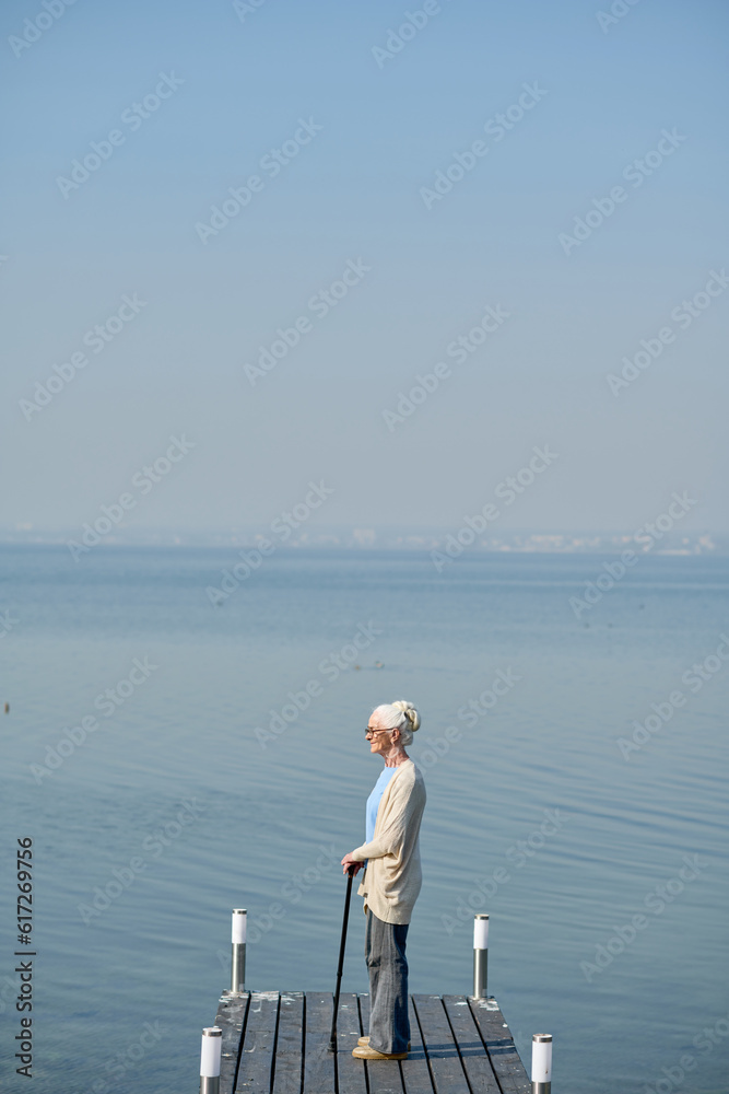 Long shot of retired woman with walking cane standing on pier by large blue lake and enjoying beautiful view while having promenade
