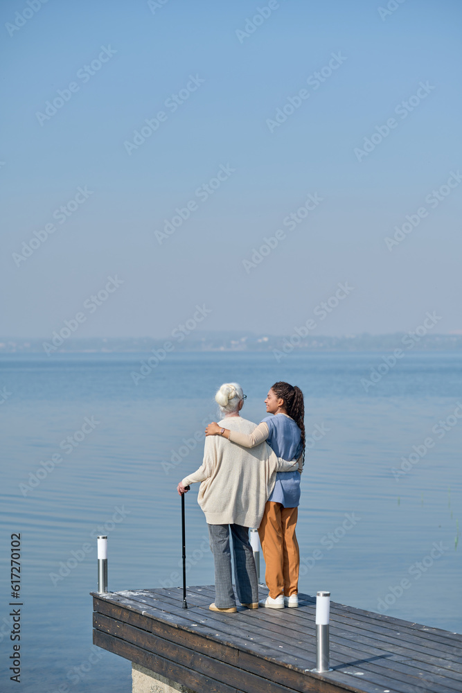 Rear view of granmother and granddaughter embracing each other while standing on pier and enjoying strolling together by waterside