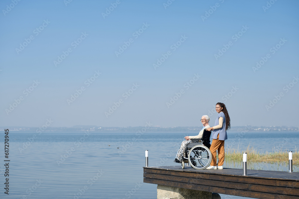 Long shot of granddaughter standing by her grandmother in wheelchair on pier while both enjoying beautiful scenery of wide blue lake
