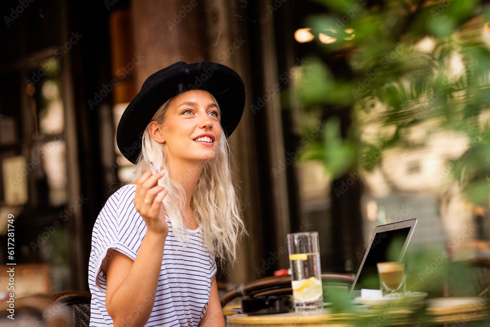 Happy young woman smoking cigarette while sitting outdoors at the cafe terrace
