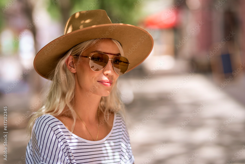Stylish blond haired woman wearing hat and sunglasses while standing on the street