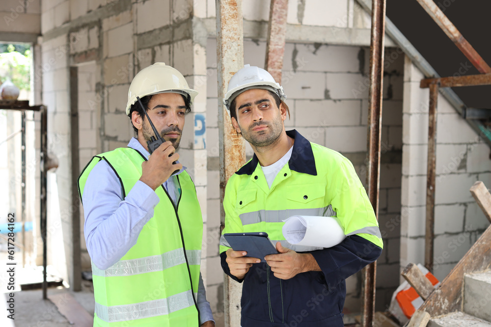 Two technician civil engineer or specialist inspector discussing, brainstorm and planing work with tablet, blueprint and walkie talkie radio together at Industrial building site. Construction concept Two technician civil engineer or specialist inspector discussing, brainstorm and planing work with tablet, blueprint and walkie talkie radio together at Industrial building site. Construction concept