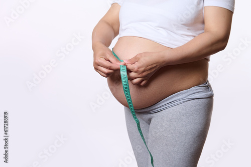 Closeup Gravid pregnant woman, expectant mother measuring her big tummy in the third trimester with a tape, isolated white studio background. Pregnancy. Maternity. Female health and gynecology concept