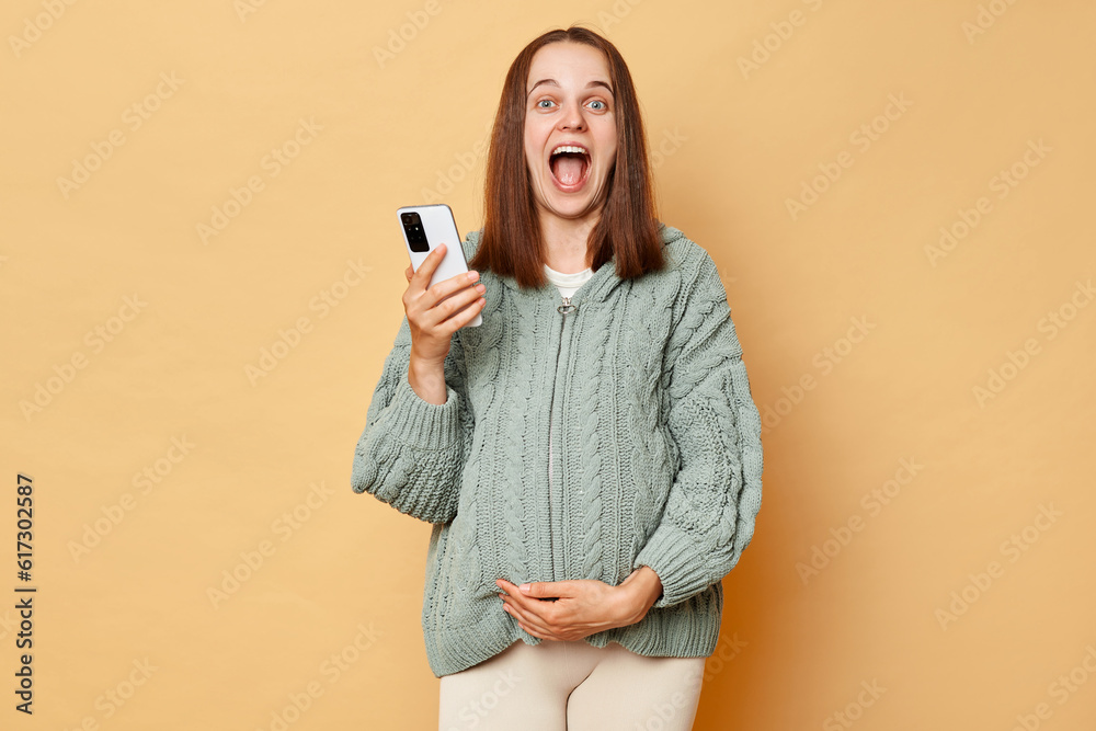 Surprised amazed pregnant woman wearing warm knitted sweater touching belly posing over beige background using cell phone looking at camera screaming rejoicing hearing good news. Surprised amazed pregnant woman wearing warm knitted sweater touching belly posing over beige background using cell phone looking at camera screaming rejoicing hearing good news.