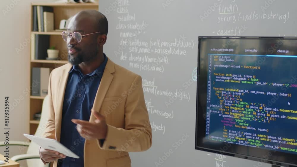Medium close up of male African American teacher standing in front of blackboard in classroom giving lecture on programming at daytimeMedium close up of male African American teacher standing in front