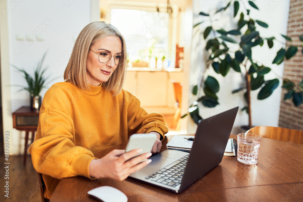 Fototapeta premium Portrait of adult woman using mobile phone while sitting in living room with laptop