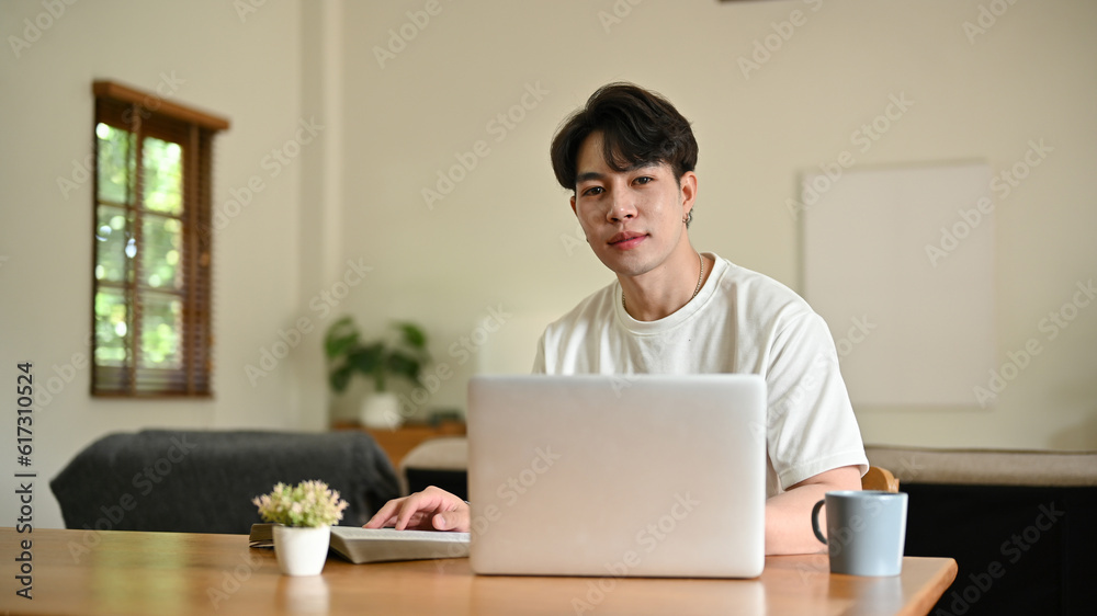 A handsome young Asian man sits at a table with his laptop in his minimalist living room. A handsome young Asian man sits at a table with his laptop in his minimalist living room.