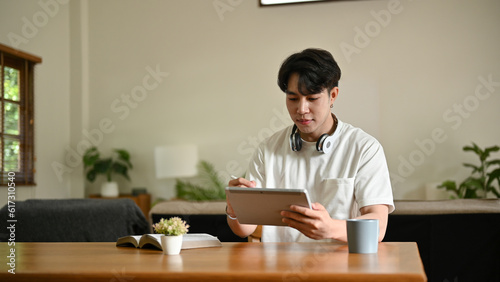 A handsome young Asian man using his digital tablet at a table in the living room.