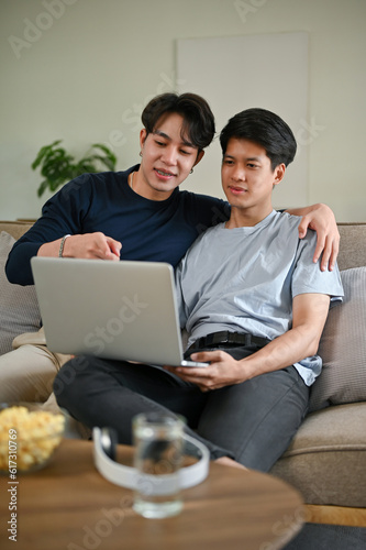 A happy Asian gay man enjoys watching a movie through a laptop with his boyfriend on a couch