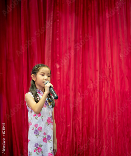 The Asian kid girl sing a song on stage at her school activity day, dress in Qipao style, red curtain background