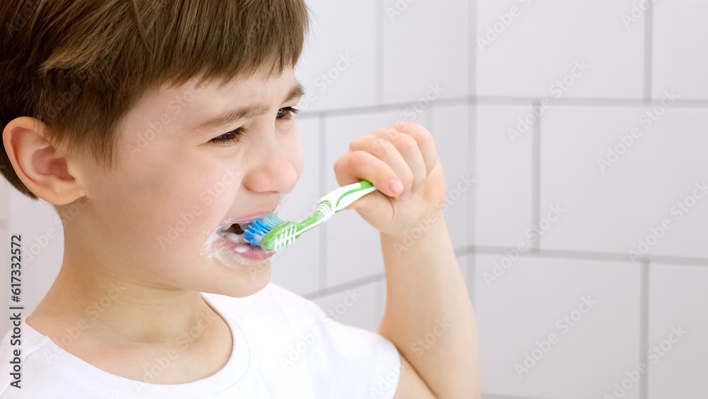 A cute little child brushes teeth. Green toothbrush. Daily morning hygiene procedures in a white bathroom. Portrait of beautiful caucasian 6 year old boy. Teeth brushing concept. Copy space. Close-up A cute little child brushes teeth. Green toothbrush. Daily morning hygiene procedures in a white bathroom. Portrait of beautiful caucasian 6 year old boy. Teeth brushing concept. Copy space. Close-up