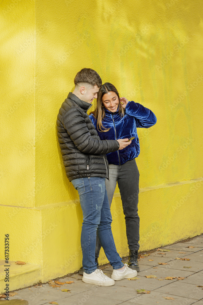 Young couple look at a mobile phone with a yellow wall in the background.