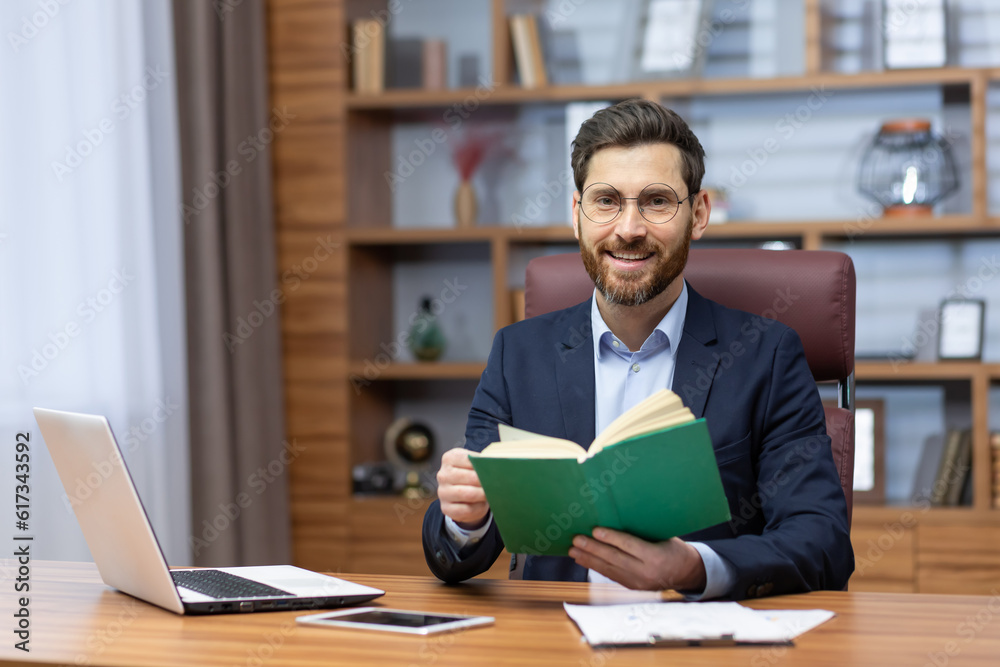 Obraz premium Joyful businessman in home office resting on break between work, mature man reading book, portrait of smiling boss holding book smiling and looking at camera.