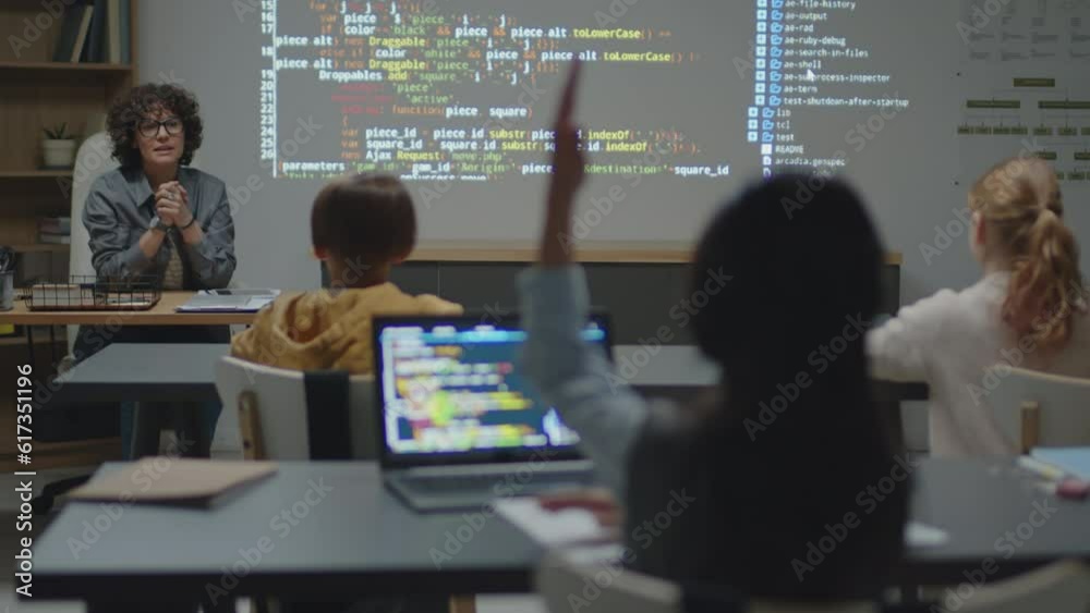 Over-the-shoulder full shot of Caucasian female teacher sitting at desk in classroom asking student raising hand to answer during programming lesson