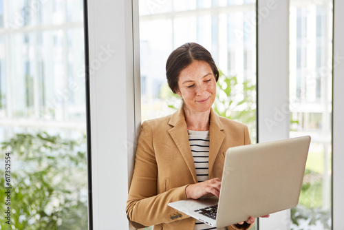 Smiling female entrepreneur using laptop while leaning on window in office lounge