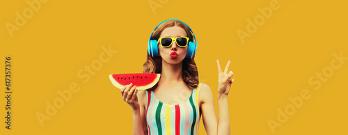 Summer portrait of stylish woman in headphones listening to music blowing her lips sends kiss with fresh juicy slice of watermelon on yellow background