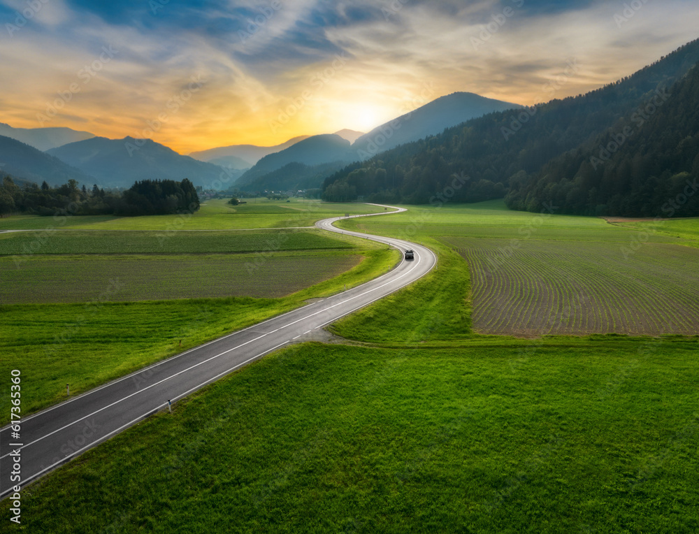 Top drone view of beautiful landscape with alpine mountains, paved highway, hills, fields green grass. Aerial view of beautiful curved rural road in green meadows at sunset in summer in Slovenia