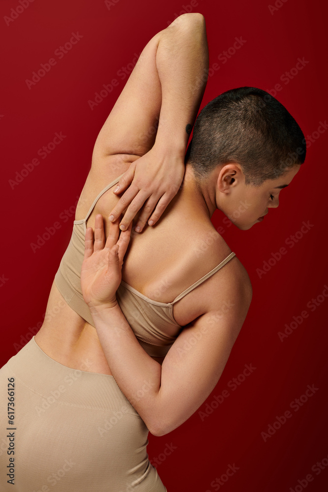 Naklejka premium body positive, tattooed young woman with short hair posing with hands behind back on burgundy background, dark red, curvy fashion, comfortable in skin, female underwear