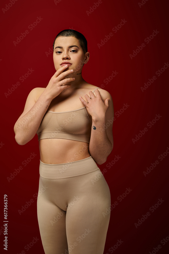Fototapeta premium body love, young tattooed woman in beige underwear posing on red background, body positivity, curvy fashion, comfortable in skin, curvy model, generation z, self love, looking at camera