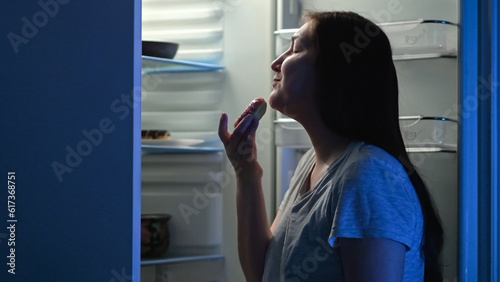 Woman opens refrigerator and looks at tempting donuts with chocolate cream at night. Brunette lady wants to eat tasty donuts during midnight photo