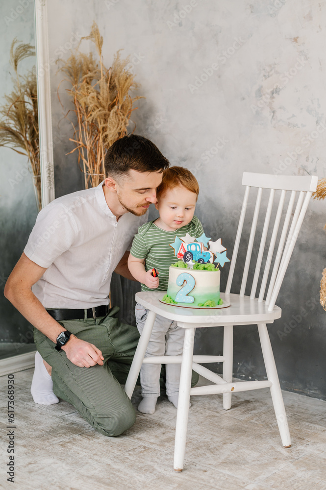 Naklejka premium Dad congratulate boy on birthday in studio. Father hugging son. Happy family blowing candles on cake and having fun celebrating birthday party 2 years old. Eat cake. Сhild closes eyes and makes wish.
