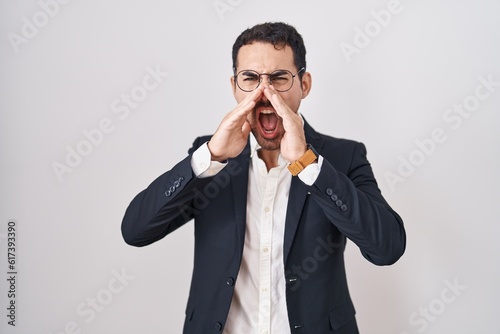 Handsome business hispanic man standing over white background shouting angry out loud with hands over mouth