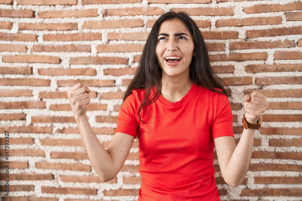 Young teenager girl standing over bricks wall angry and mad screaming frustrated and furious, shouting with anger. rage and aggressive concept. Young teenager girl standing over bricks wall angry and mad screaming frustrated and furious, shouting with anger. rage and aggressive concept.