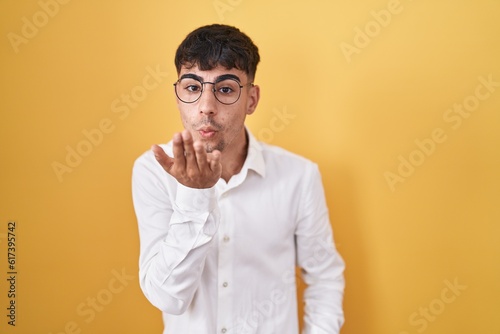 Young hispanic man standing over yellow background looking at the camera blowing a kiss with hand on air being lovely and sexy. love expression. © Krakenimages.com
