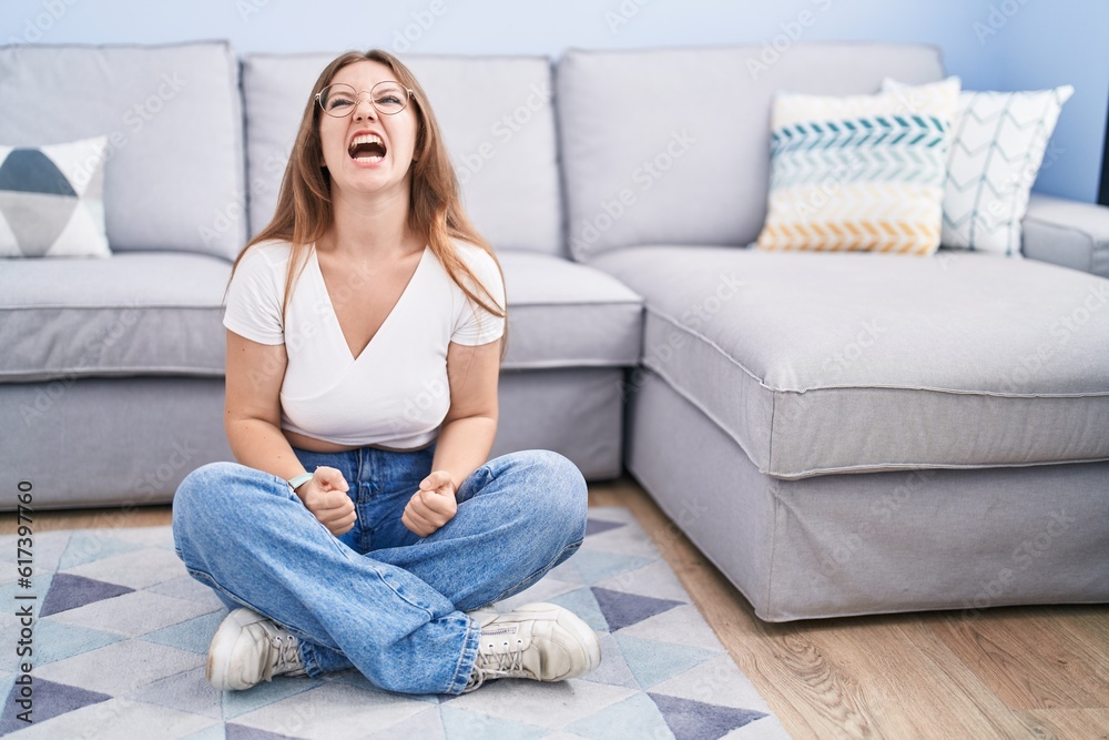 Young caucasian woman sitting on the floor at the living room angry and mad screaming frustrated and furious, shouting with anger. rage and aggressive concept.