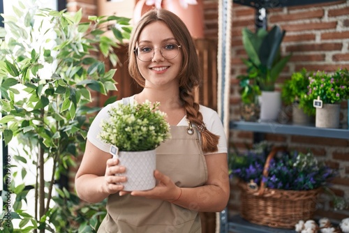 Young caucasian woman working at florist shop holding plant looking positive and happy standing and smiling with a confident smile showing teeth