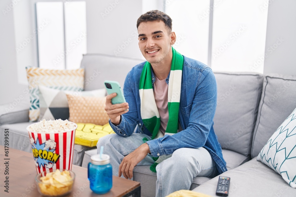Young hispanic man soccer fan using smartphone sitting on sofa at home Young hispanic man soccer fan using smartphone sitting on sofa at home