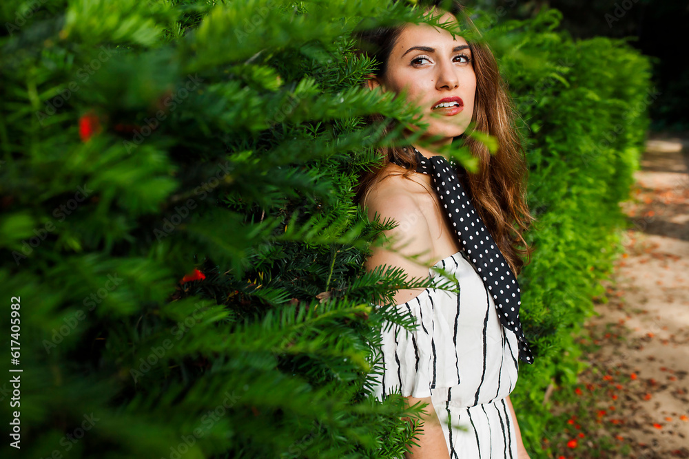 Side view of beautiful female with red lipstick in white off-the-shoulder dress pressing back to pine tree and looking at camera with slightly open mouth. Side view of beautiful female with red lipstick in white off-the-shoulder dress pressing back to pine tree and looking at camera with slightly open mouth.