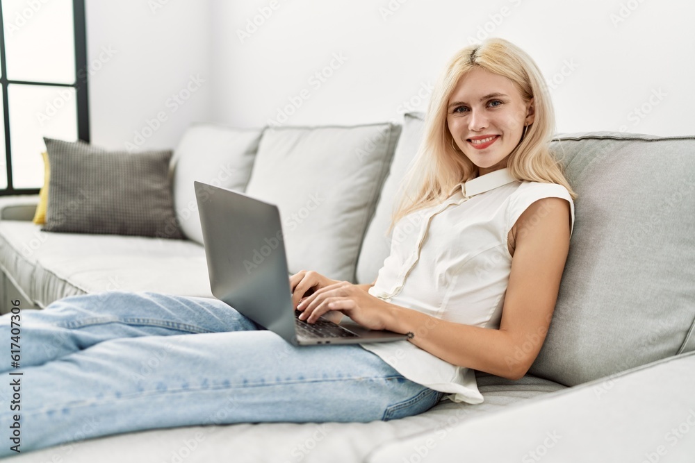 Young blonde woman using laptop sitting on sofa at home