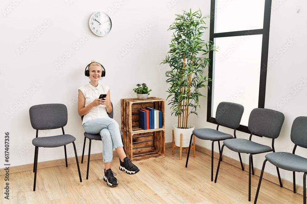 Young blonde woman using smartphone and headphones sitting on chair at waiting room