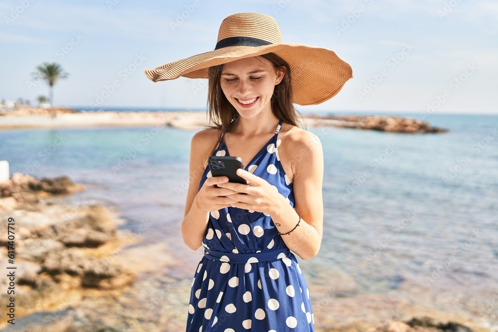 Young blonde woman tourist wearing summer hat using smartphone at beach