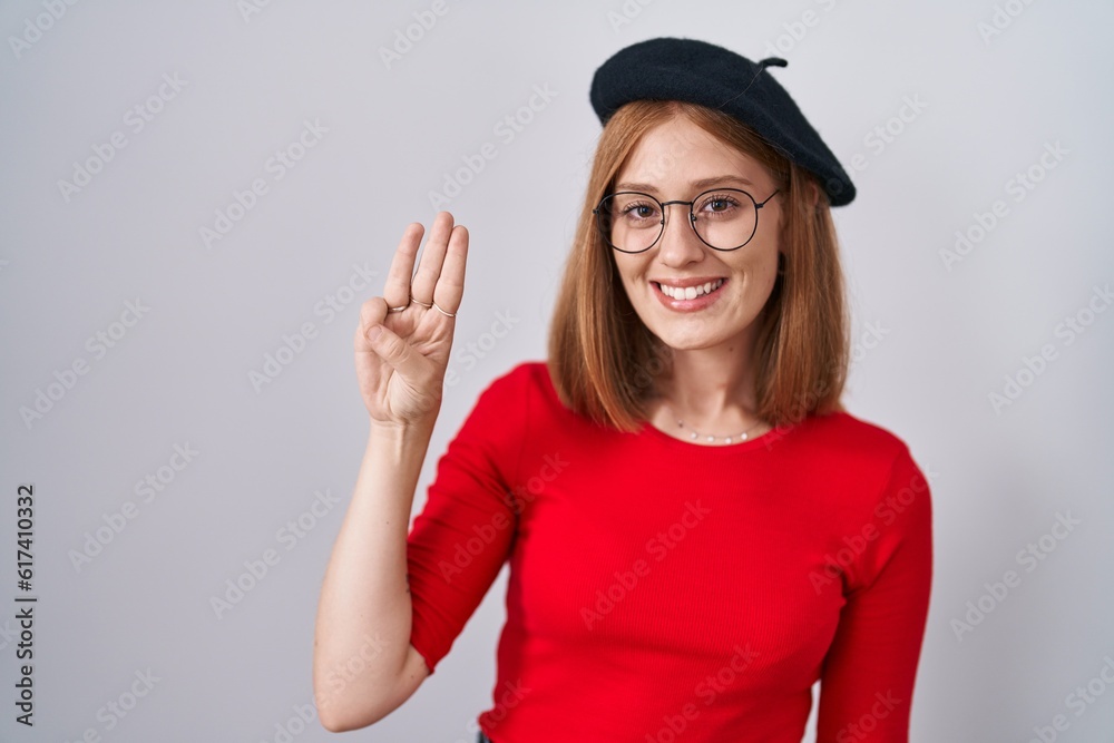 Young redhead woman standing wearing glasses and beret showing and pointing up with fingers number three while smiling confident and happy. Young redhead woman standing wearing glasses and beret showing and pointing up with fingers number three while smiling confident and happy.